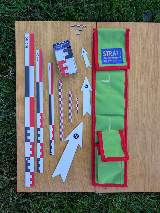 Open CANOMA 50 cm canvas archaeological photography scale kit with green red roll-up case, metric scales and white north arrows on wood and grass