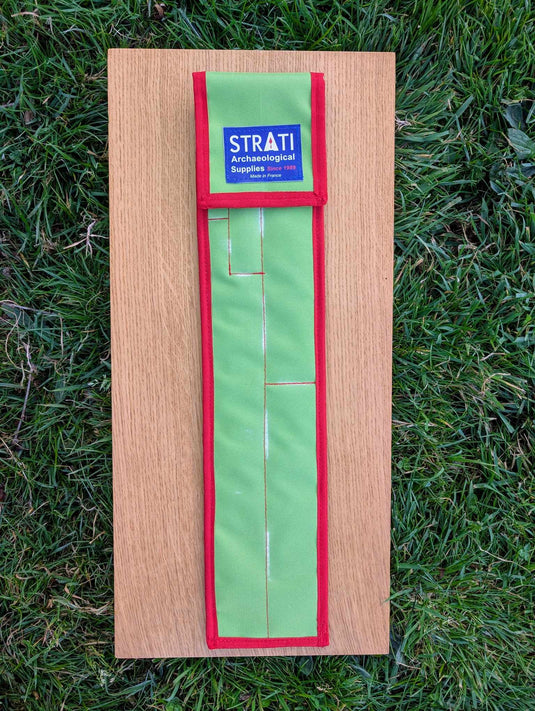 Closed green and red STRATI CASE carrying case for archaeological photo scales on a wooden board over grass