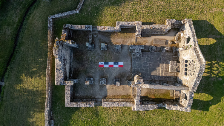 Aerial drone photo of stone ruin foundations in a grassy field with a 5m CANVAS photo scale bar in red-white-black placed at the center for size reference.