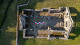 Aerial drone photo of stone ruin foundations in a grassy field with a 5m CANVAS photo scale bar in red-white-black placed at the center for size reference.