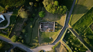 Chargez l'image dans la visionneuse de la galerie, Top-down drone view of rectangular stone ruins surrounded by fields, roads and hedges. CANVAS 5m archaeological scale visible inside the structure.

