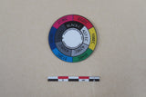 Small circular Strati SCD colorimeter disk with multicolor and greyscale Pantone segments on beige background