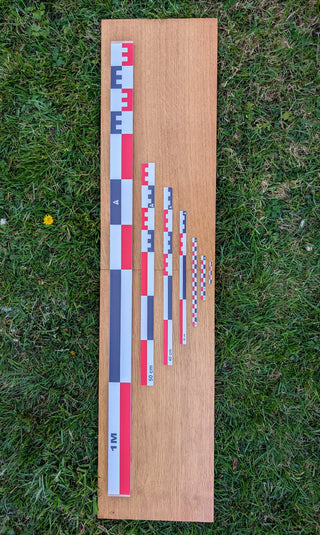 Chargez l'image dans la visionneuse de la galerie, Seven flat archaeological photo scales (5cm to 1m) in red-white-black, arranged on a wooden board on grass. Complete graduated set. Made in France.
