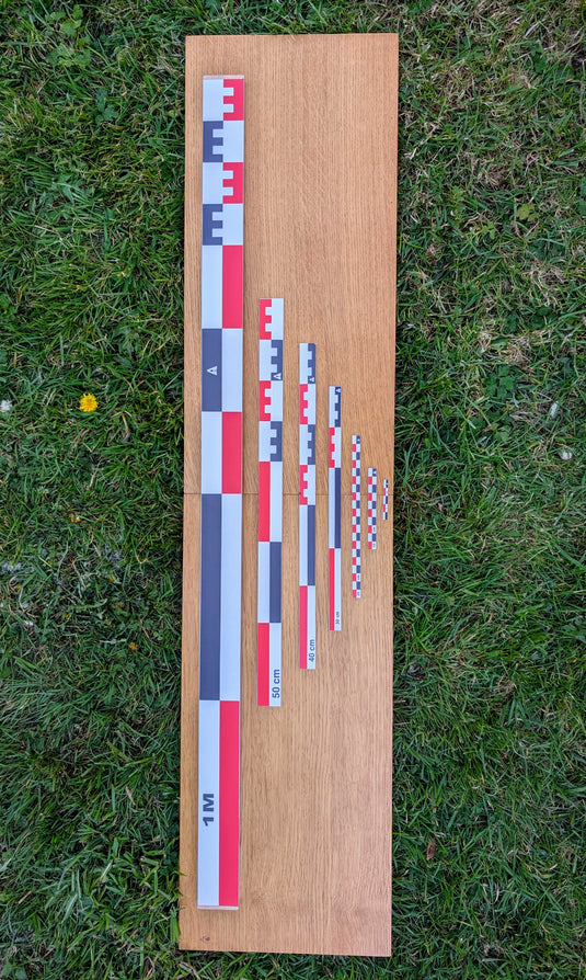 Seven flat archaeological photo scales (5cm to 1m) in red-white-black, arranged on a wooden board on grass. Complete graduated set. Made in France.