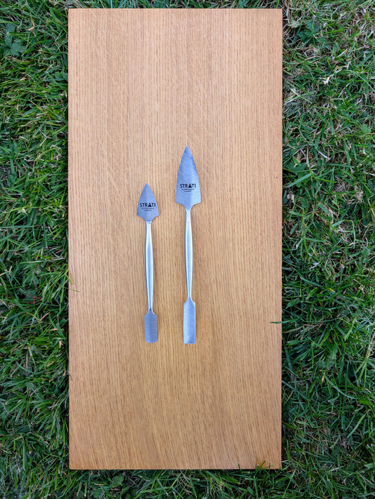 Two forged stainless steel spatulas, small and large, with pointed and flat ends on wood over grass
