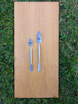 Two forged stainless steel spatulas, small and large, with pointed and flat ends on wood over grass