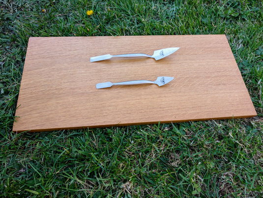 Top profile view of two forged stainless steel spatulas on light wood board over green grass
