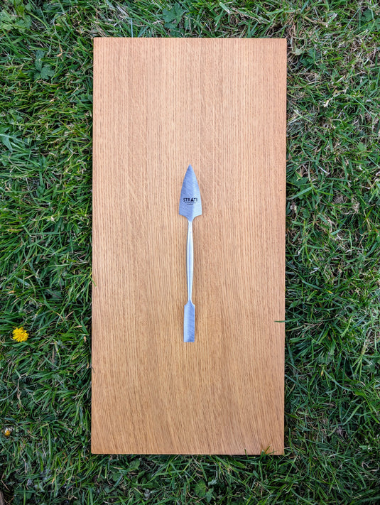 Forged stainless steel dual-ended spatula with brushed finish on light oak board over green grass