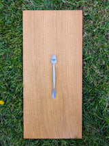 Small forged stainless steel dual-ended spatula with brushed finish on light oak board over grass